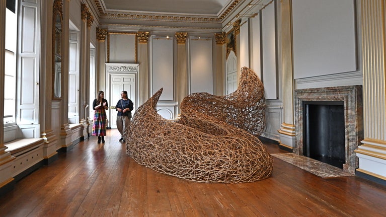 Two people looking at a large willow sculpture in a grand historic room with wooden floors and golden pillars